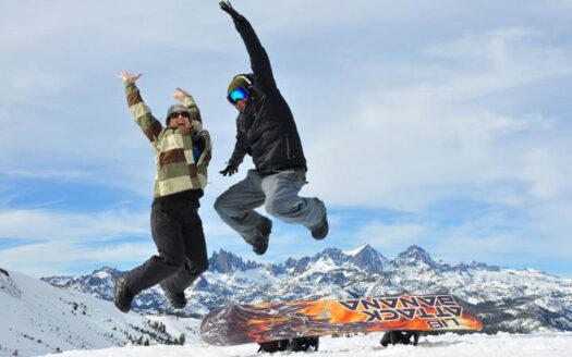 Two people jump joyfully in the snowy mountains of Mammoth Lakes, California, with a snowboard resting in the foreground and jagged peaks in the background.
